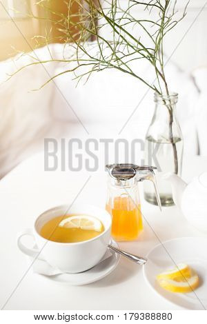 Cup of hot green tea with lemon on white table, fresh lemon and honey - closeup shot. Sun is shining through the window, warm feeling. Relaxation and enjoying live concept