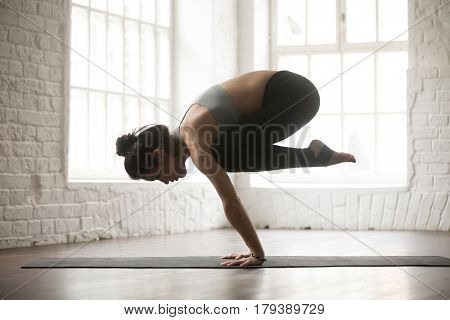 Young attractive yogi woman practicing yoga concept, standing in Bakasana exercise, Crane pose, working out, wearing black sportswear bra and pants, full length, white loft studio background