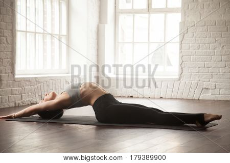 Young attractive yogi woman practicing yoga concept, stretching in Matsyasana exercise, Fish pose, working out, wearing black sportswear, full length, white loft studio background