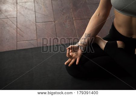 Young yogi woman practicing yoga concept, sitting on black mat in studio or home in Padmasana exercise, Lotus pose, working out, meditating, close up of hand with tattoo, copy space floor background