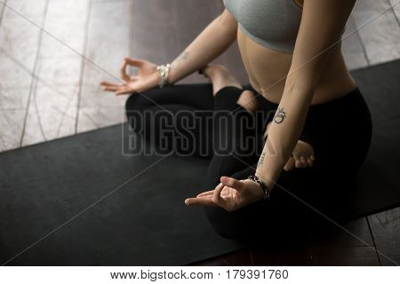 Midsection close up of young yogi woman practicing yoga on black mat, sitting in Padmasana exercise, Lotus pose, working out, wearing sportswear bra and pants, studio background. Meditation session
