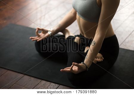 Close up of young woman with tattoo and nice wrist bracelets practicing yoga, sitting in Padmasana exercise, Lotus pose with mudra, working out on black mat, wearing sportswear, studio background