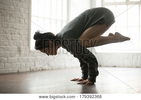 Young attractive yogi woman practicing yoga concept, standing in Bakasana exercise, Crane pose, working out, wearing grey oversized sweater, full length silhouette on white loft studio background