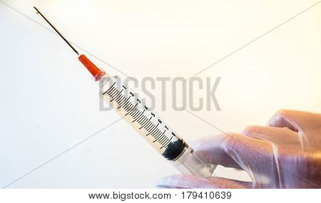 A full syringe drips while a latex-gloved hand prepares to give an injection against a white background