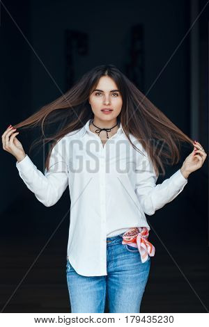 Portrait of pensive white Caucasian brunette young beautiful woman touching her long hair wearing white shirt and blue jeans standing on black dark background