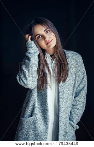 Portrait of smiling white Caucasian brunette young beautiful girl woman model with dark hair and brown eyes in white shirt and grey long coat looking in camera on black background