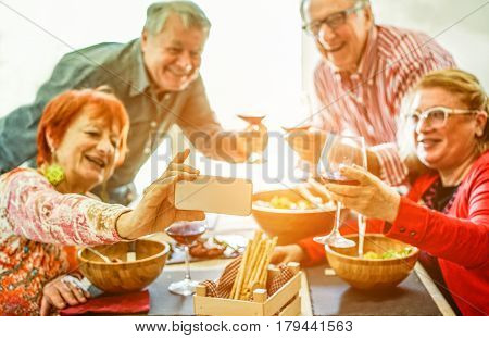Senior couples taking selfie at home toasting red wine and eating lunch meal - Old friends having fun with new trends technology - Joyful elderly lifestyle concept - Focus on phone hand - Warm filter