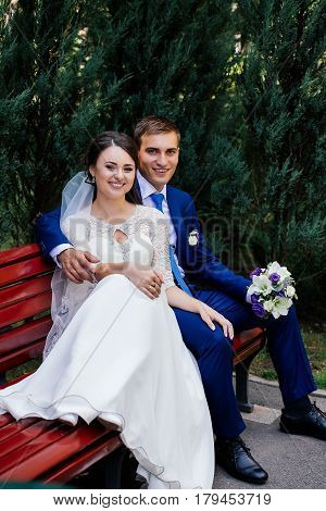The bride and groom sit on a bench. The groom hugs the bride
