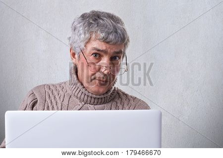 A confident mature businessman wearing glasses working with a laptop while being at home looking into camera with serious expression. People lifestyle and technology concept.