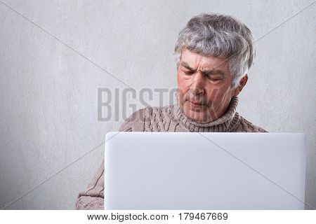 A mature man with gray hair siting in front of his laptop having dissatisfied expression and wrinkles on his face isolated over white background. People emotions technology and lifestyle concept.