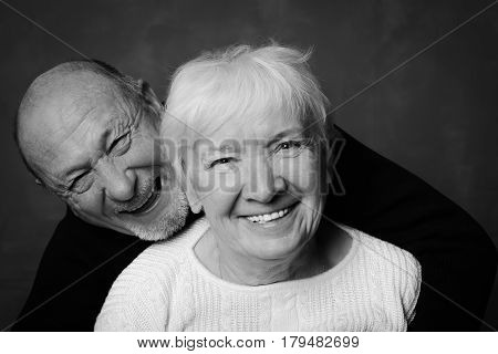 Black and white studio shot of handsome senior couple on a dark background. Man in black hugs his wife in white smiling and laughting looking at the camera. Love and devotion concept