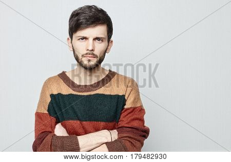 Sudio shot of young brunette attractive guy gazing at the camera with suspicion. Caucasian man in vintage sweater crosses his hands and staring forward.