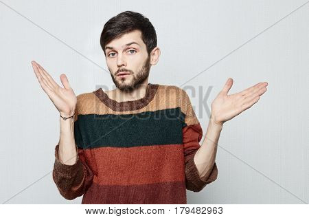 Studio shot of young bearded Causasian man in old fashioned colorful sweater spreads his hands in amazement to the sides. In surprise he looks forward. Human emotional concept.