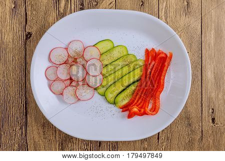 Fresh sliced vegetables lie on a plate on a wooden background. Vegetables seasoned with spices: salt, black and red ground pepper, as well as lightly sprinkled with vegetable oil.