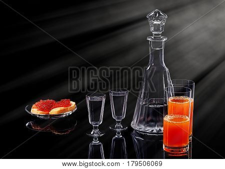 Decanter and two shot glasses with ice vodka, two salmon red caviar sandwiches on a glass plate and two glasses with multifruit juice on a black acrylic surface with reflection
