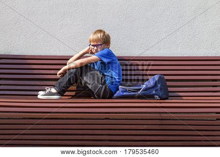 Tired schoolboy with glasses sitting on the bench after school at sunny day