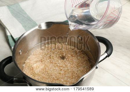 Pouring water into saucepan with brown rice on kitchen table