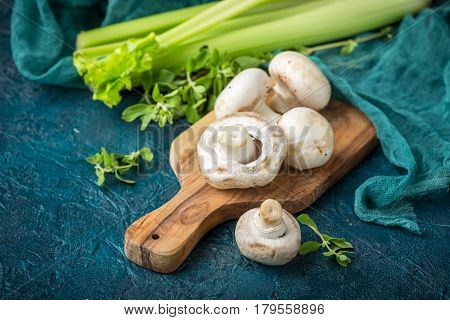 Fresh mushrooms champignons on a wooden cutting board parsley and stalks of celery on a dark blue background