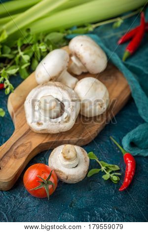 Fresh mushrooms champignons on a wooden cutting board parsley greens celery stalks cherry tomatoes and hot red chili pepper on a dark blue background
