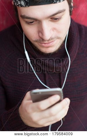 handsome young man with a beard wearing a woolen sweater and a baseball cap with a smartphone and wearing headphones looks at the smartphone chatting