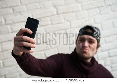 grimacing young man in a cap and sweater makes selfie with smartphone on brick wall background