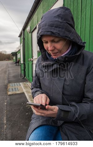 Authentic woman in winter jacket using mobile phone to type sms message outdoors while sitting on the bench on a cold day