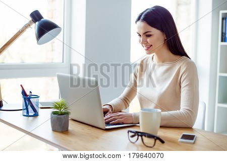Young Smart Smiling Beautiful Woman Sitting At The Table And Typing Letter To Her Besiness Colleague