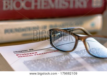 Eye glasses on top of an open book with Protozoa Medical Parasitology in focus with other life science text books in the background out of focus