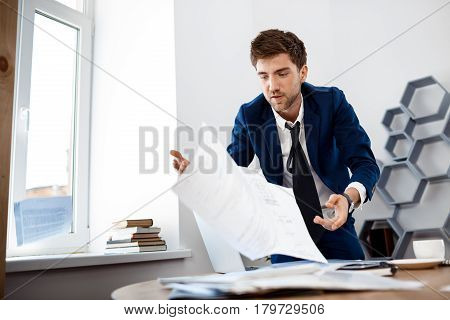 Absentminded young businessman in suit rummaging in papers at workplace, office background.