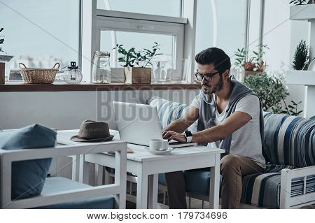 Concentraiting on project. Handsome young modern man in eyewear using computer while sitting on the sofa
