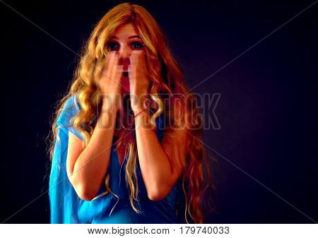 Frightened woman screaming with fear at dark halloween nightat horror of what she saw. Girl in long blue evening dress with tousled hair bawl covered her face on black background.