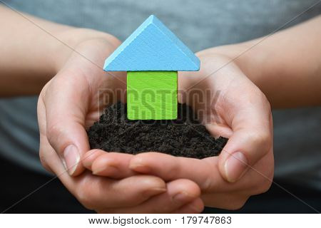 Female hands holding a piece of land with wooden house. Ecological home, construction and real estate concept