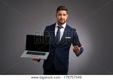 An elegant handsome young man holding and showing the screen of a laptop while looking uncomprehendingly in to the camera