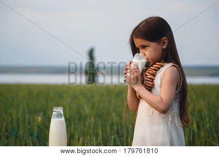 Happy small girl in a white dress drinking milk in a green field