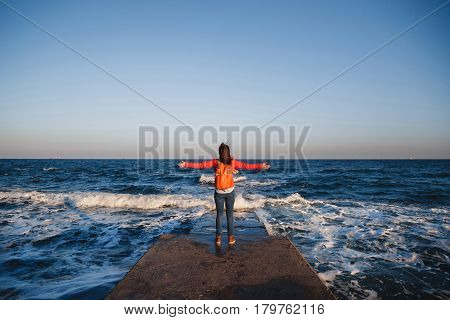 Young happy girl with backpack standing on a seaside breathing fresh air raising arms enjoying the view