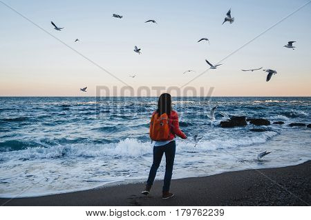Young happy girl with orange backpack feeding seagulls on a seaside evening time