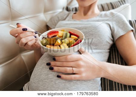 Closeup of pregnant young woman sitting on sofa and eating fruit salad at home