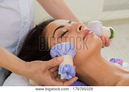 Herbal ball face massage in ayurveda spa. Female massagist with young woman in wellness center. Healthcare therapy to beautiful indian girl in beauty parlor, side view of face with eyes closed