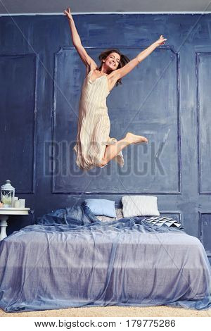 Low angle of a cheerful woman jumping on the bed with outspread arms in the bedroom. Girl having fun. Bedroom interior 