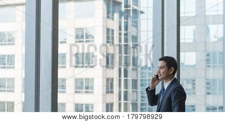 Mature and confident business executive looking looking out of large windows at a view of the city below from the top floor of an office building while talking on his mobile phone - panoramic banner