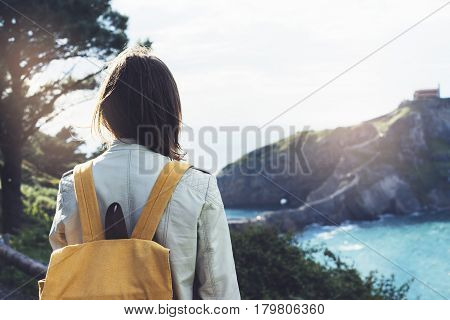 Hipster young girl with backpack enjoying sunset on seascape on peak mountain. Tourist traveler on background valley landscape view mockup. Hiker looking sunlight ocean in trip holiday in basque island