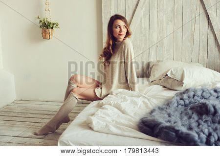 young woman in knitted cardigan and warm socks wake up in the morning in cozy scandinavian bedroom and sitting on bed with white bedlinen. Casual lifestyle in modern interior