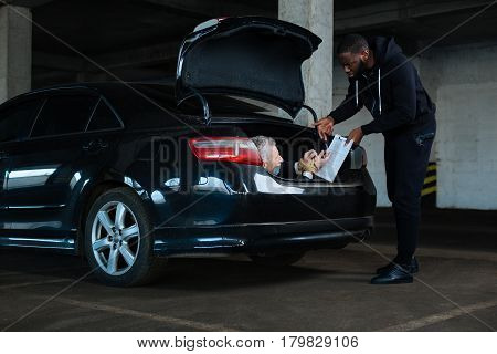 I will do everything. Sad scared senior man lying in the car boot and signing the document while being forced to do this