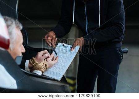 Sign this. Unhappy sad serious man having his hands tied and being forced to sign a document while being kidnapped