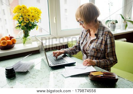 The elderly woman works behind the laptop. On a table various receipts and accounts lie. Modern pensioners use modern technologies.