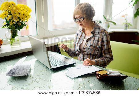 The elderly woman works behind the laptop. On a table various receipts and accounts lie. Modern pensioners use modern technologies.