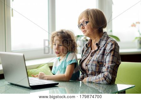 The grandmother and the grandson with interest look at the laptop screen. The elderly woman and the boy sit next. Modern technologies are available to people of all age.