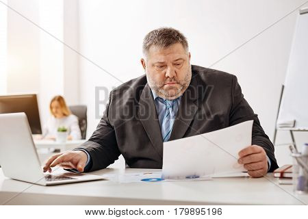 Paying attention. Tired scrupulous office clerk working at his desk and analyzing sales report while typing the data on his computer