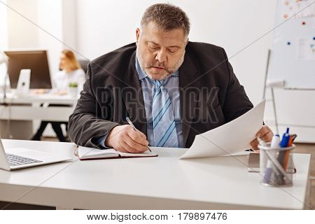 Thorough analysis. Diligent scrupulous puffy man fulfilling his work while analyzing a report and making some notes