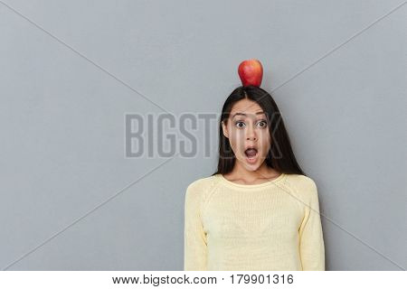 Amazed astonished young woman with red apple on her head standing and shouting over grey background
